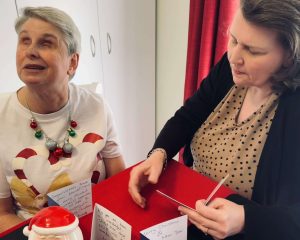 A photo of Cathy seated at a table with a staff member who is reading out Christmas cards to her. Cathy is wearing a Christmas t-shirt and ball-ball necklace.