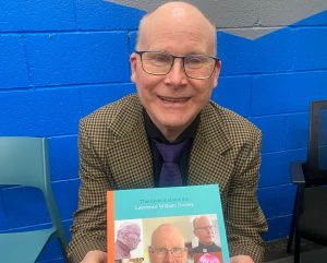 A photo of Lawrence sitting down holding his book 'This book is about me' smiling and he is wearing glasses.