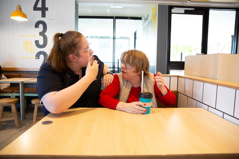 Two women sitting in a cafe. The woman on the left is signing in Auslan to the woman on the right.