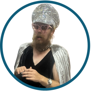 Jasper is dressed in a sparkling, silver costume, wearing an ornate, jewel‑covered headpiece and a metallic silver cape over a black sparkly sleeveless top. He has a studded choker around his neck, and his hands are positioned in front of them, with dark nail polish visible.