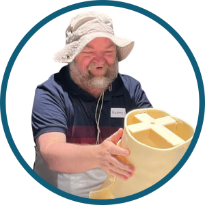 Rodney is smiling wearing a wide‑brimmed sun hat and a navy polo shirt with a name tag. He holds a large yellow plastic object with cross‑shaped openings.