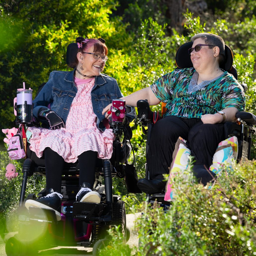 Margie and Rachael sit next to each other in their wheelchairs, surrounded by green bushes. They are looking at each other and smiling.