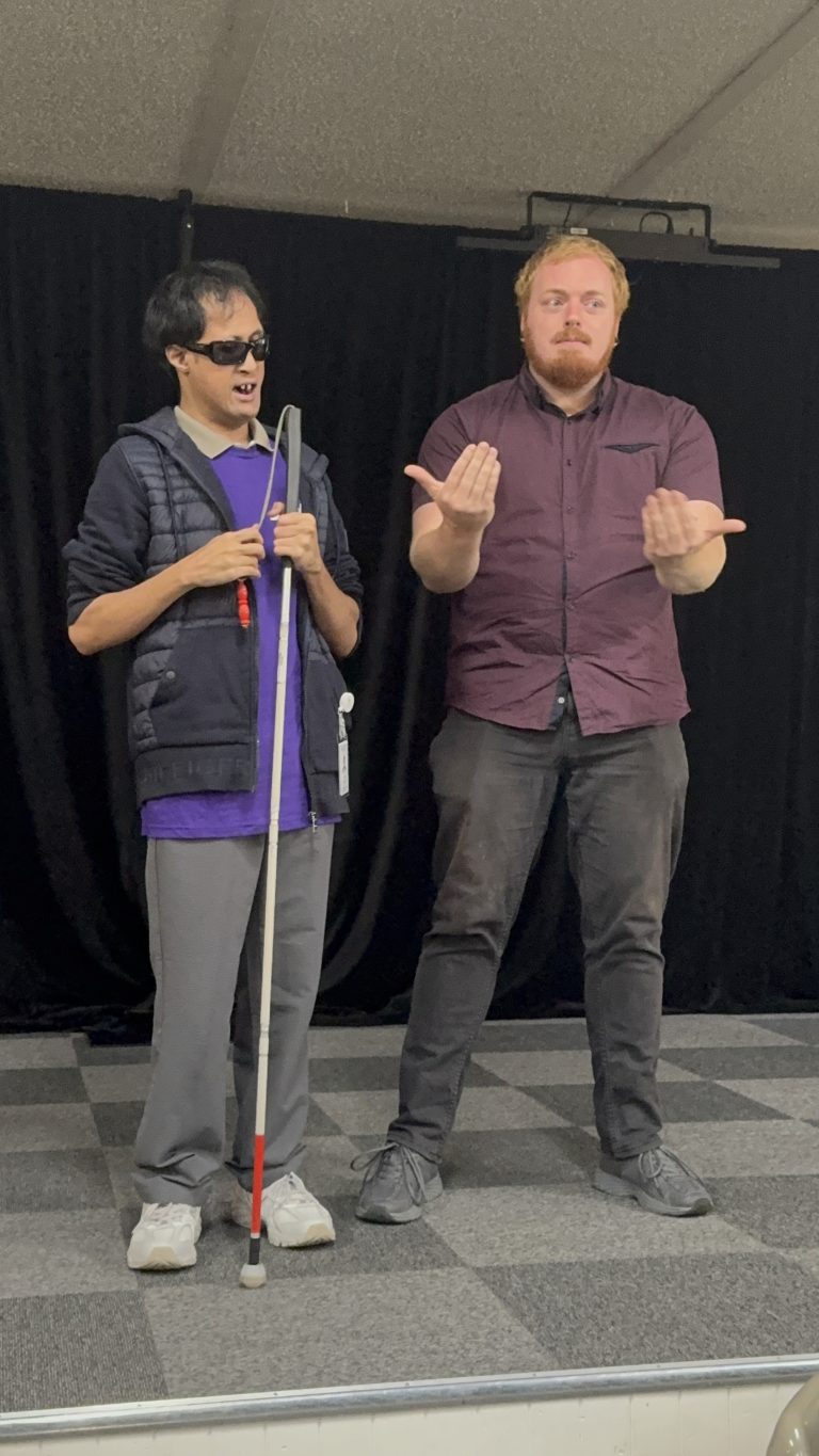 Alex standing on stage presenting at the Deafblind Camp 2026 and to his left is his interpreter