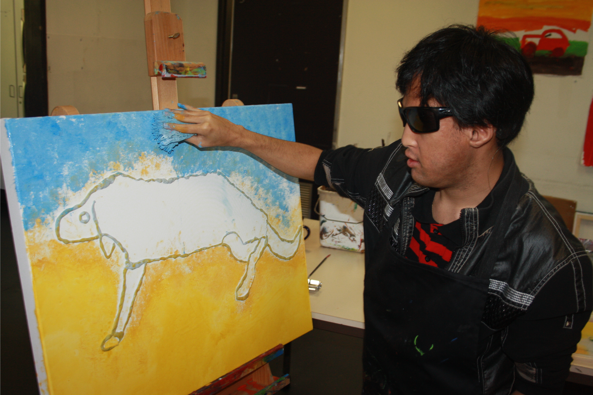 Alex painting on a canvas. He is painting an animal with a blue background that represents the sky.