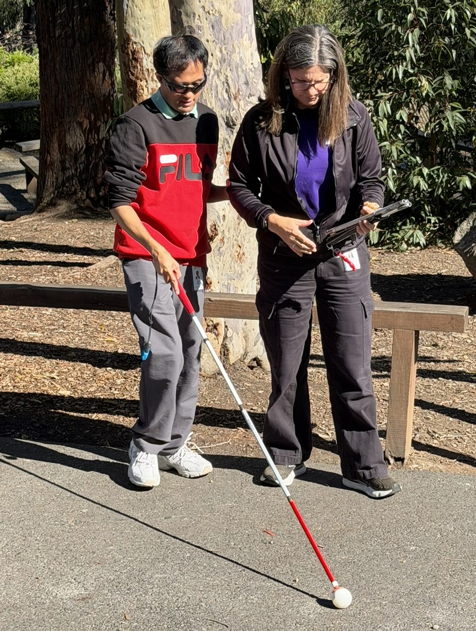 Alex is walking with his cane outside and is being supported by his Able Australia CommGuide