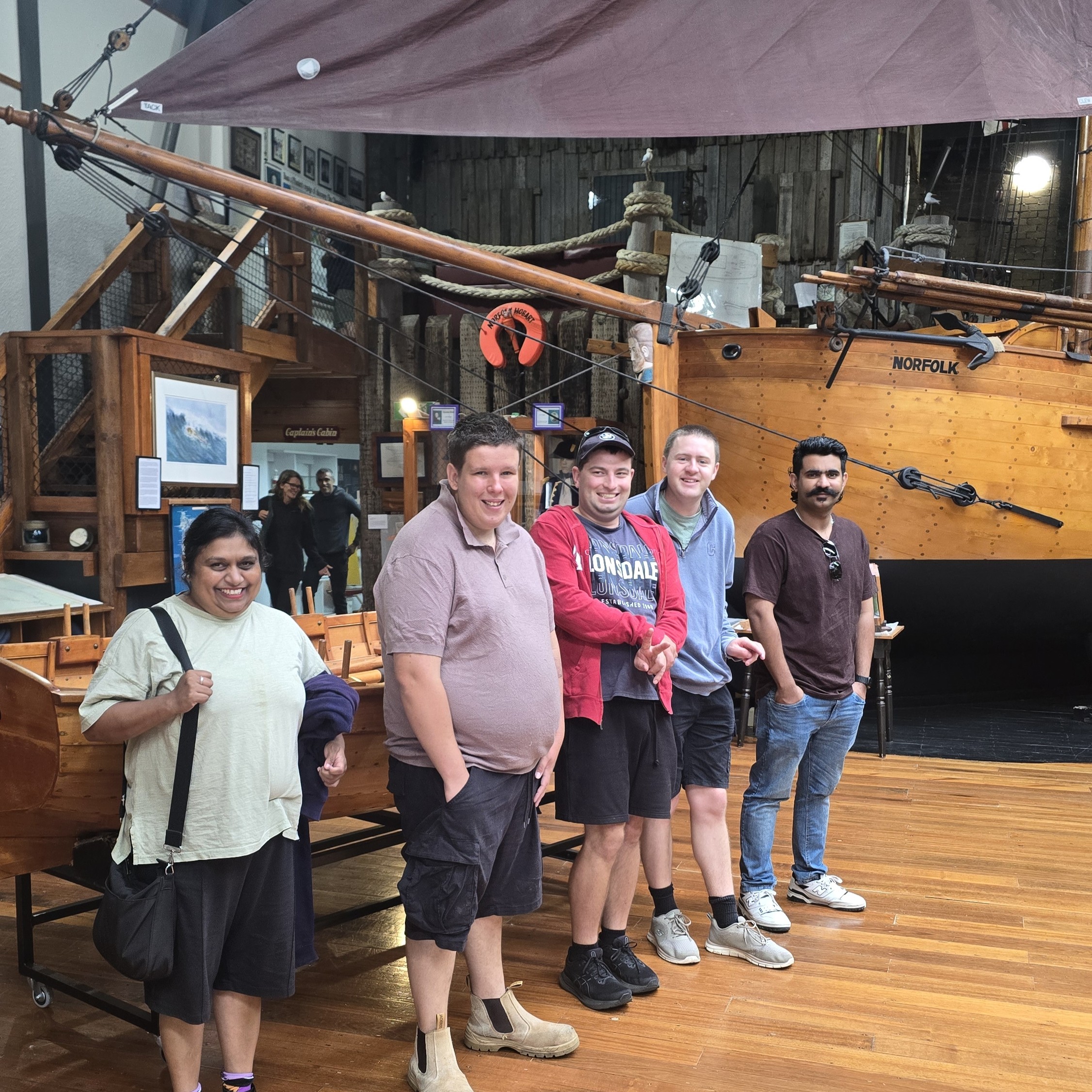 Five people stand inside a maritime museum beside a wooden boat display, with ropes, rigging and the word “NORFOLK” visible on the hull.