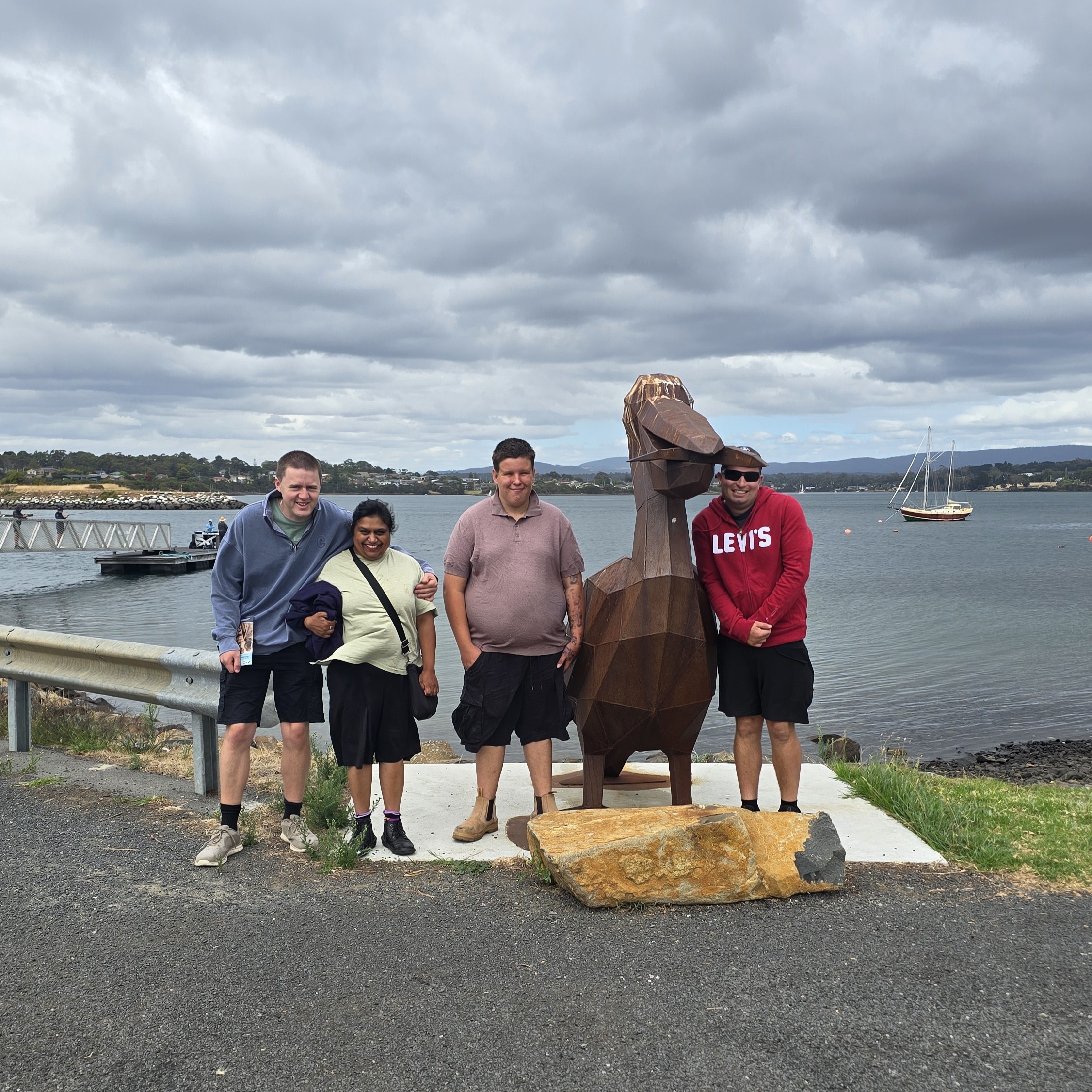 Four people stand beside a metal pelican sculpture holding a fish, near a waterfront with a sailboat and cloudy sky in the background.