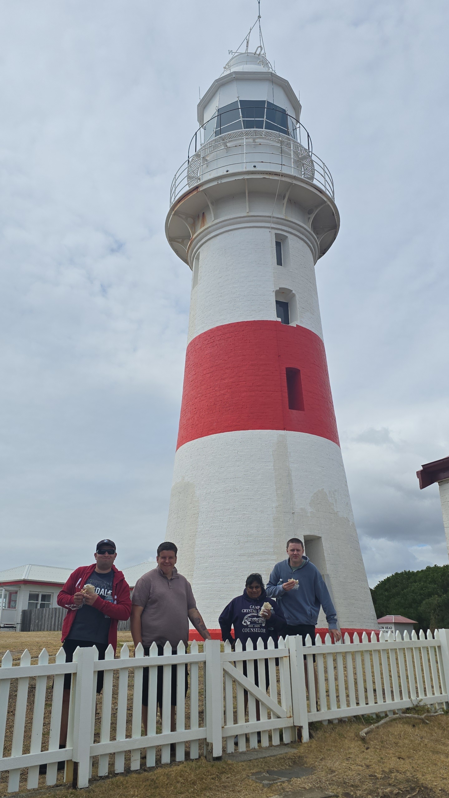 Four people stand in front of a tall red and white lighthouse behind a white picket fence, with cloudy skies overhead.