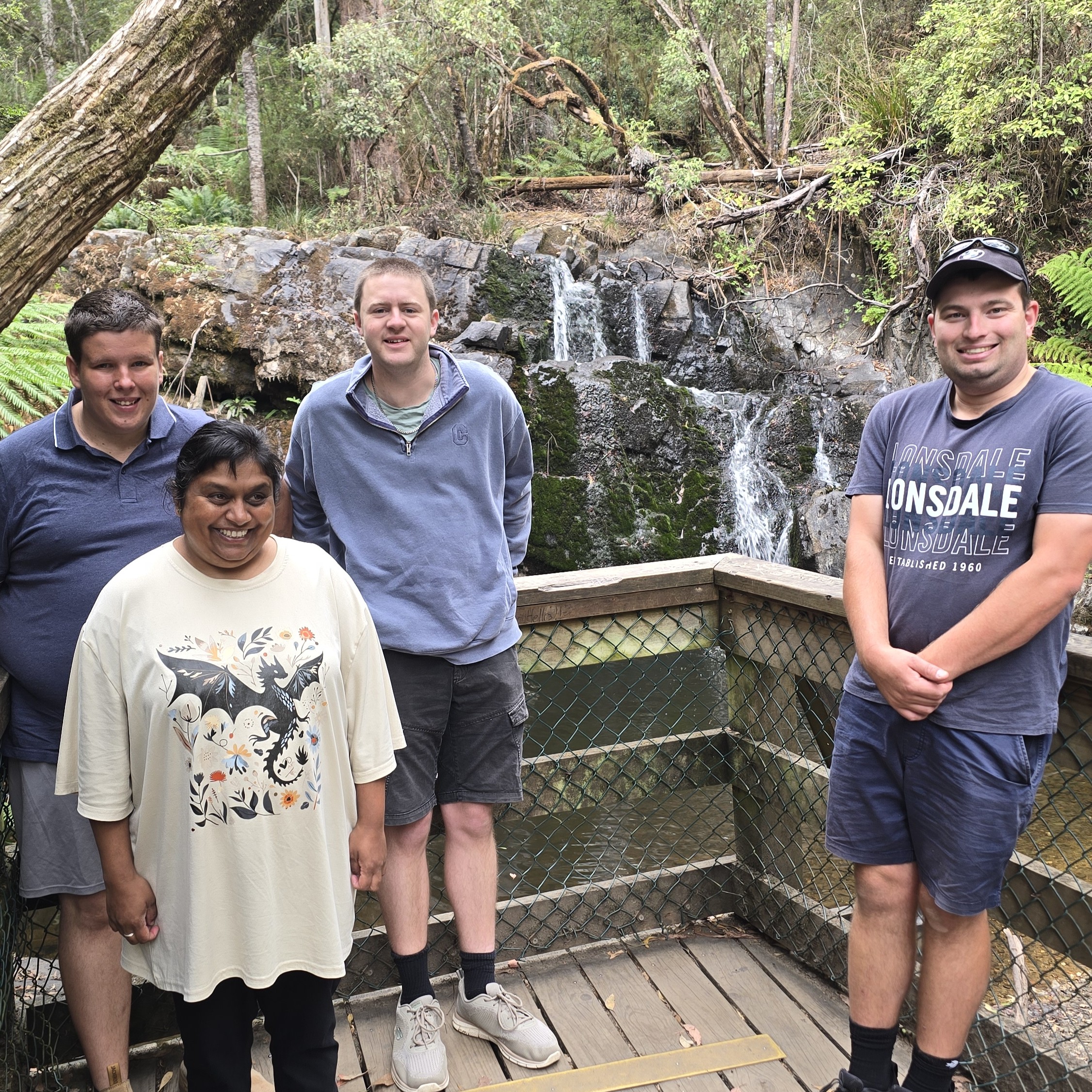 Four people stand on a wooden viewing platform in front of a small waterfall and rocky creek, surrounded by forest.