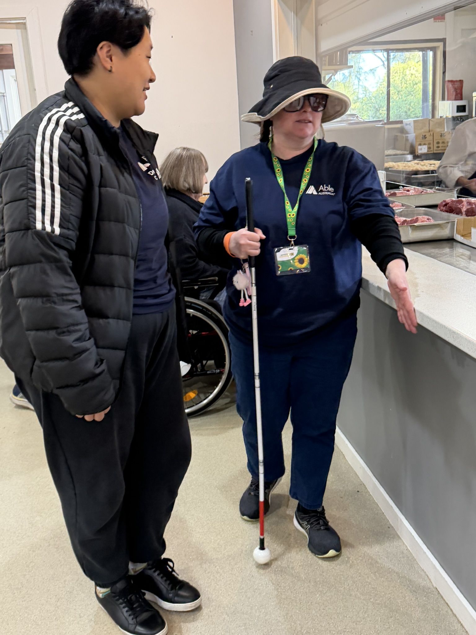 Two adults stand side by side inside a kitchen or food preparation area. One person stands on the right, holding a white cane upright in one hand and extending the other hand forward toward a bench, as if demonstrating or locating the edge. This person wears a wide-brimmed hat, dark sunglasses, a navy shirt with an “Able Australia” logo, and a lanyard with an ID card. The second person stands on the left, facing the person with the cane, wearing a black jacket with white stripes down the sleeves, a dark shirt, and black pants. Behind them is a counter with trays of raw meat and a staff member partially visible working behind a glass or open serving area. In the background, there is a window letting in daylight, stacked boxes, and kitchen equipment. A wheelchair is visible behind the two people, partly obscured. The space is well-lit with neutral-coloured walls, and the floor appears to be a smooth, light surface.