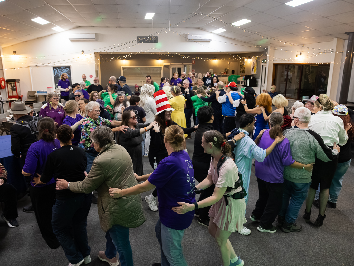A large group of adults is gathered in a bright indoor hall, arranged in a wide circle. Each person stands close behind the next, with hands gently placed on the shoulders of the person in front, forming a continuous line around the room. In the foreground, several people wear casual clothing such as jackets, jumpers, and t-shirts in colours including purple, green, black, and beige. One person near the front right wears a light pink outfit and a harness-style device with small electronic components attached around the body. Another person closer to the centre wears a tall red-and-white striped hat. The group extends across the room, creating a full circle. People are standing close together, moving or stepping in the same direction, suggesting a coordinated group activity such as dancing or a guided movement exercise. The room has light-coloured walls and a high ceiling with bright rectangular lights. Strings of small warm fairy lights are draped across the ceiling, adding a festive atmosphere. Around the edges of the space are tables, chairs, and various items, including a popcorn machine and a sign on the wall that reads “MITEY” (partially visible). In the background, more people stand and watch or join the circle. The overall environment suggests a social event or group session in a community or activity space.
