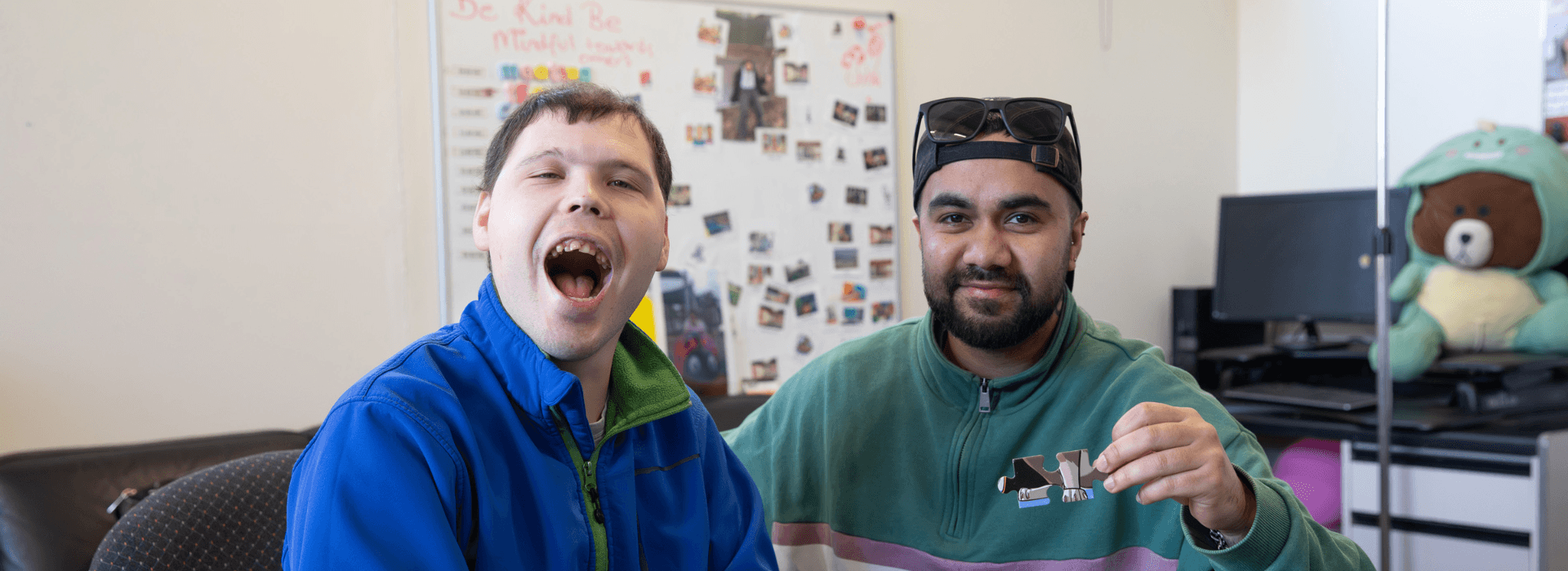 Two males sitting insdie a recreation room putting together a puzzle. The male on the right has a big smile on his face wearing a bright blue jumper. To the right is a male smiling with sunglasses on his head and green jumper